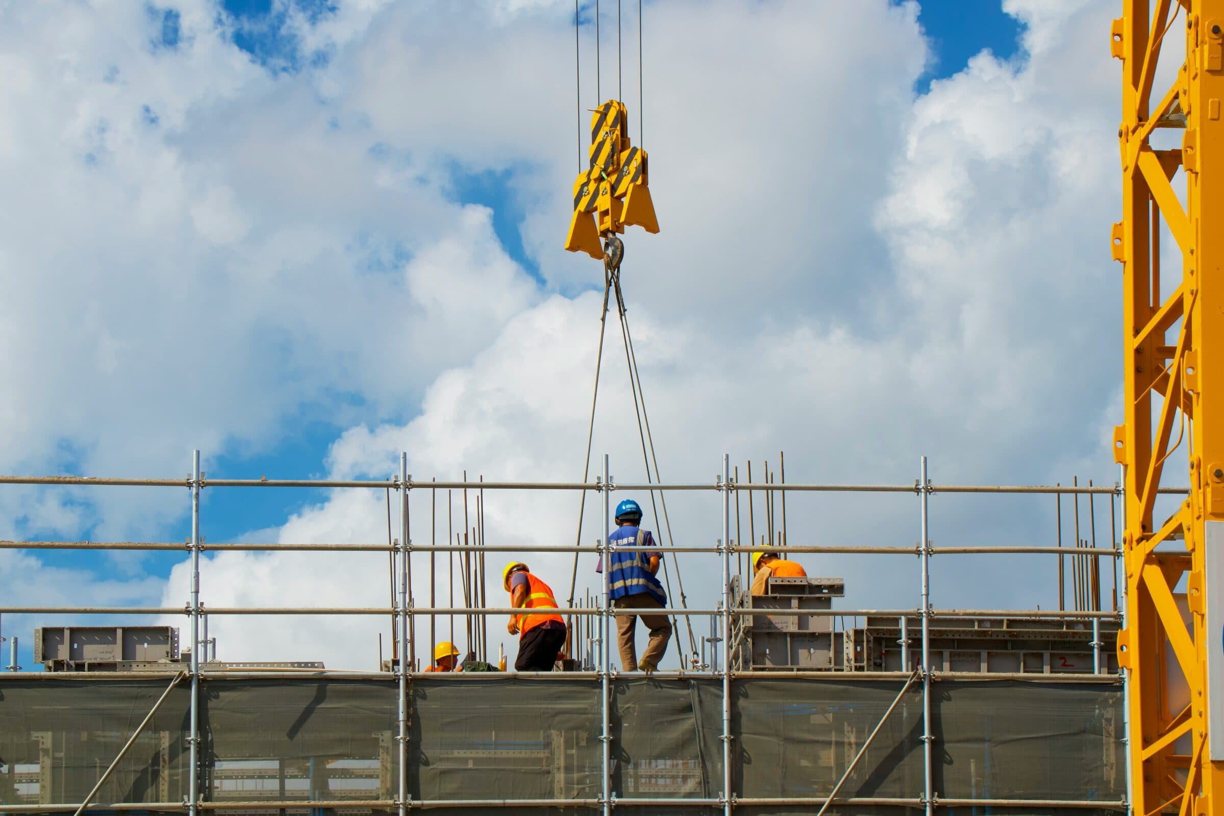 A crane hook lowering its load to four workers on a scaffolded deck, under a bright, clouded sky